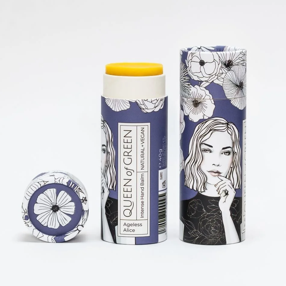 Handbalsam „Ageless Alice“ von Queen of Green in dunkelblauer, floral gestalteter Verpackung aus recyceltem Karton teilweise geöffnet mit sichtbar gelbem Balm; natürliche, vegane Handpflege im Stick.