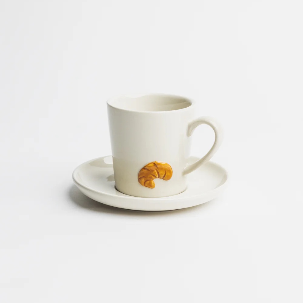Handgefertigte Espressotasse aus Steinzeug mit glänzender Glasur in naturweiß, verziert mit einem erhabenen, handillustrierten Croissant-Motiv – jedes Stück ein individuelles Unikat.